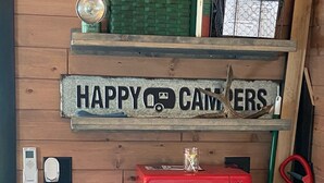 Interior d'una cabana de fusta amb un cartell que diu 'HAPPY CAMPERS', un microones vermell antic i una cistella verda a l'estant.