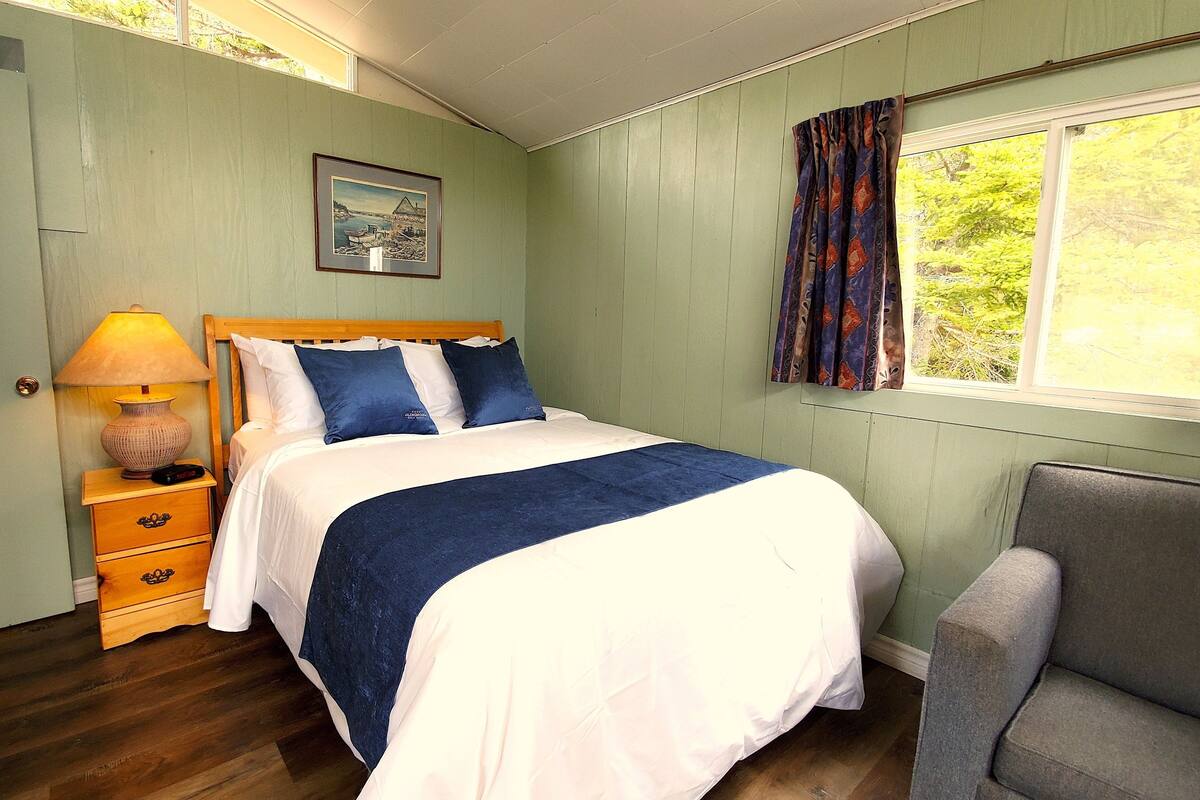 A neatly made bed with a blue and white bedspread, a bedside lamp, a wooden nightstand with drawers, a framed picture on the wall, and a window with curtains.