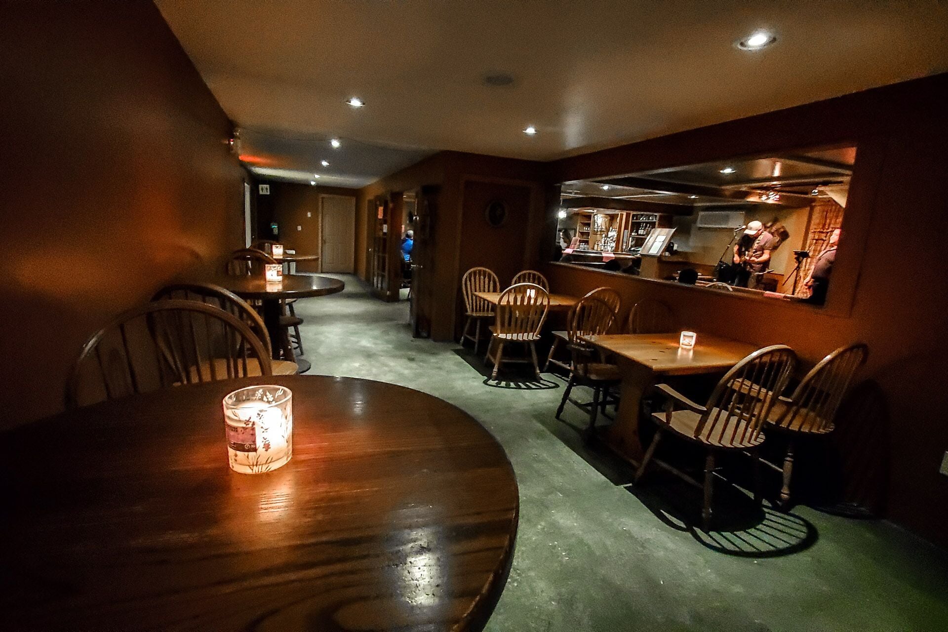 Een zwak verlicht restaurant met houten tafels en stoelen, een bar en een zichtbare keuken op de achtergrond.