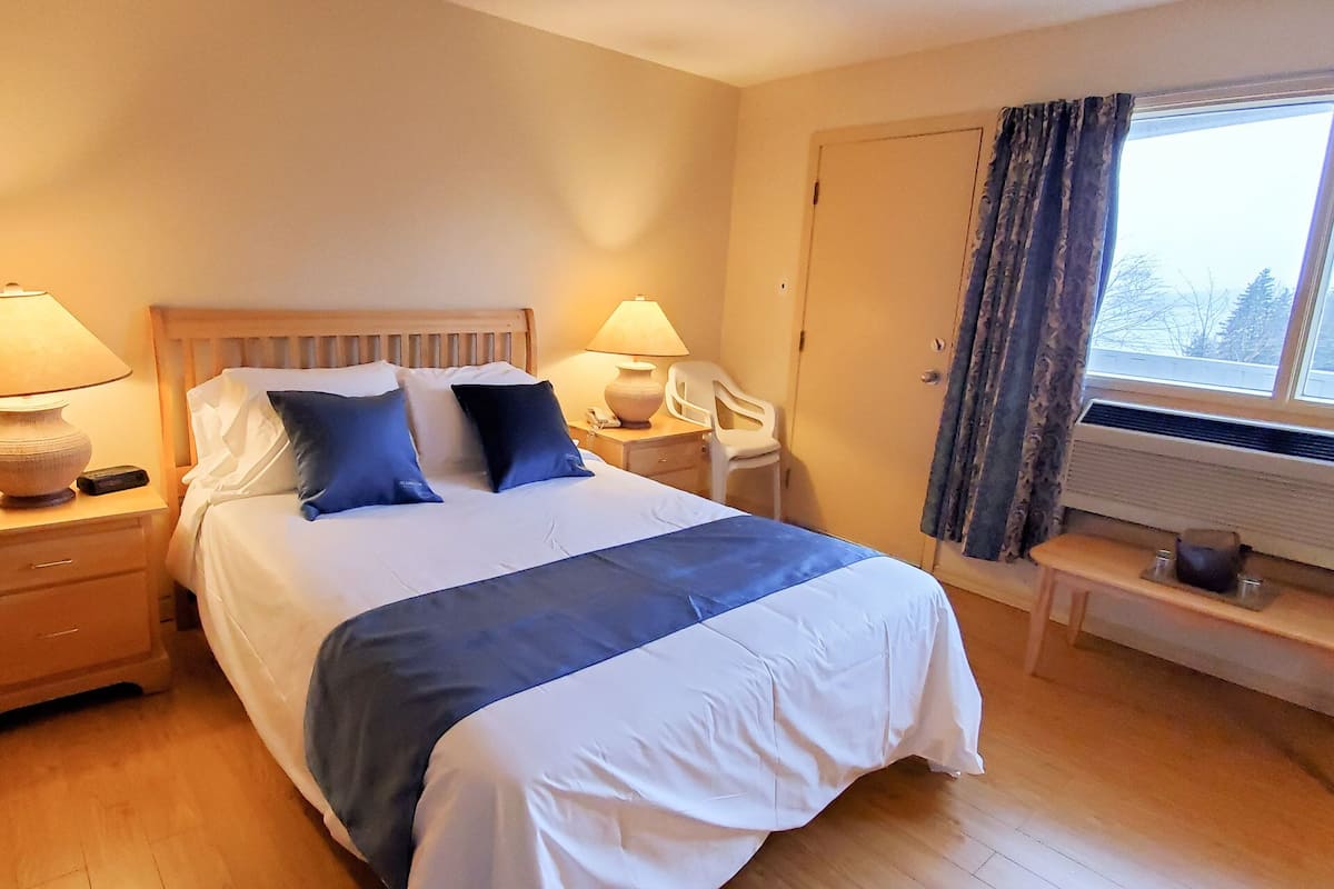 A neatly made bed with a blue and white duvet, two bedside lamps, a bedside table with a lamp, a chair, and a window with curtains.