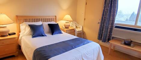 Un letto rifatto con un piumone blu e bianco, due abat-jour, un comodino con lampada, una sedia e una finestra con tende.