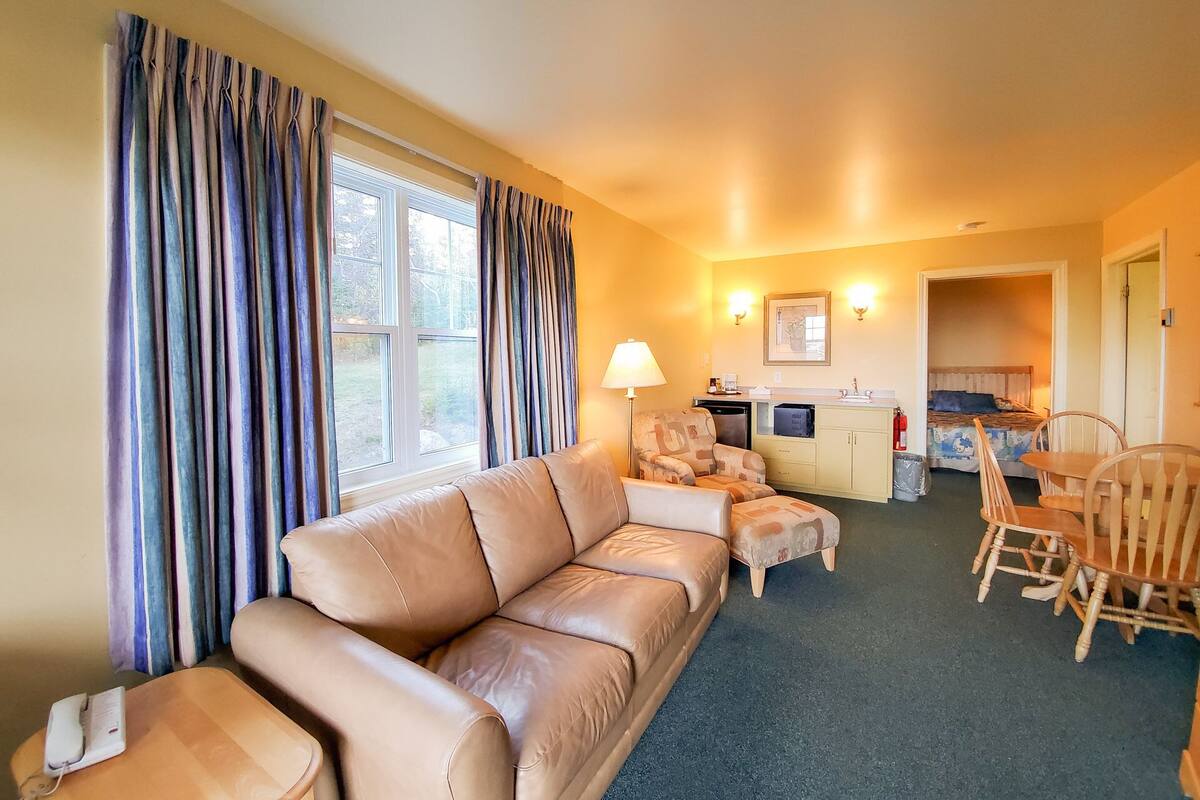 A cozy hotel room with a leather sofa, a dining table with chairs, a kitchenette, and a bed with a blue headboard.