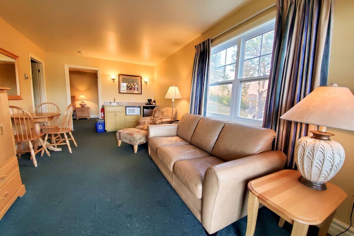 A cozy hotel room with a sofa, a dining table with chairs, a kitchenette, and a large window with curtains.