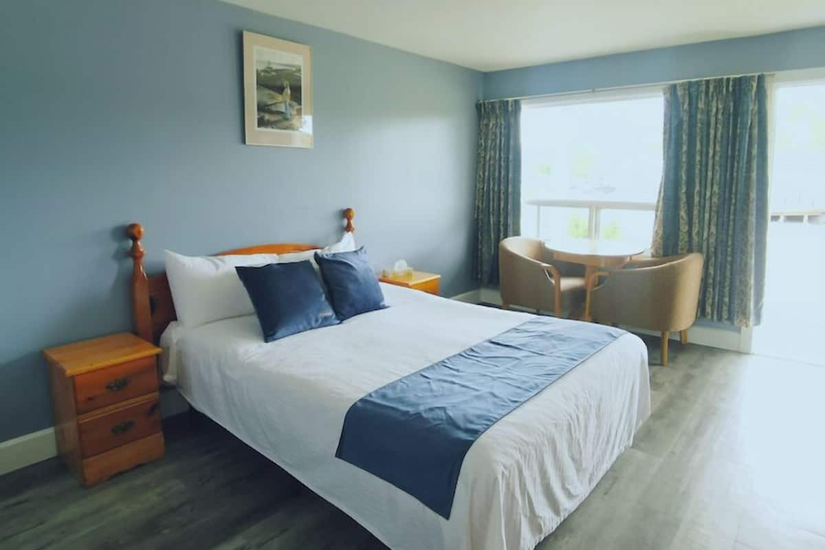 A hotel room with a bed, a wooden nightstand, a chair, a small table, and a window with curtains.