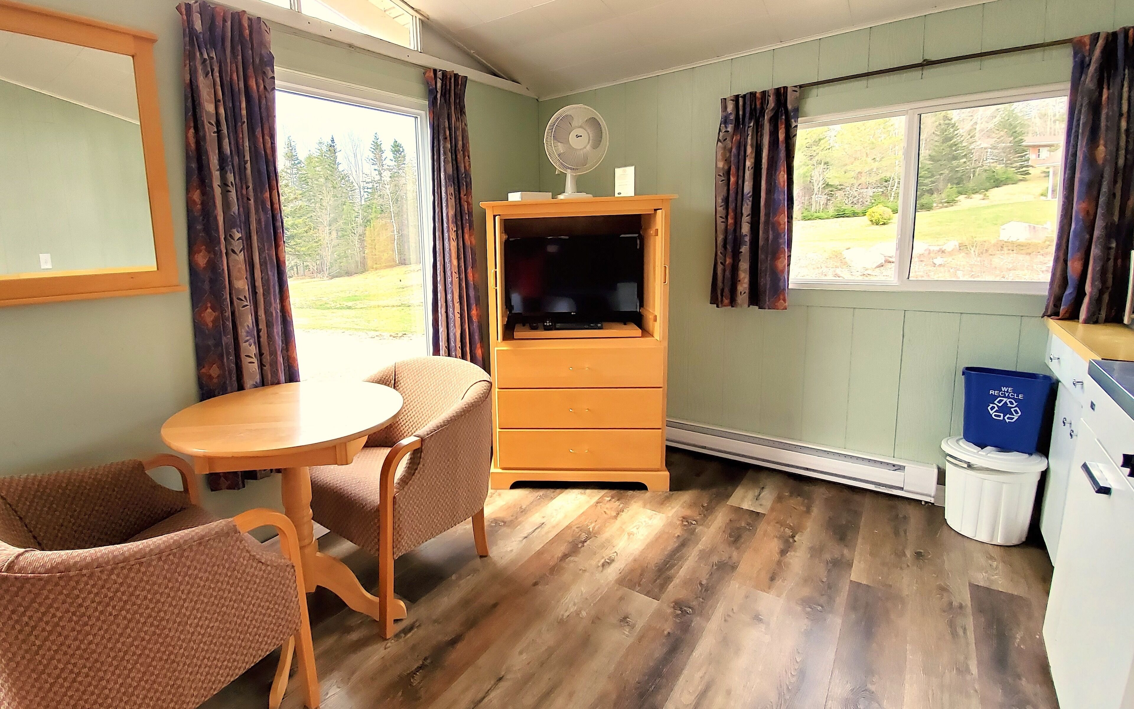 Une pièce avec une table et des chaises en bois, un téléviseur posé sur un meuble en bois, une fenêtre avec des rideaux et une poubelle de recyclage.