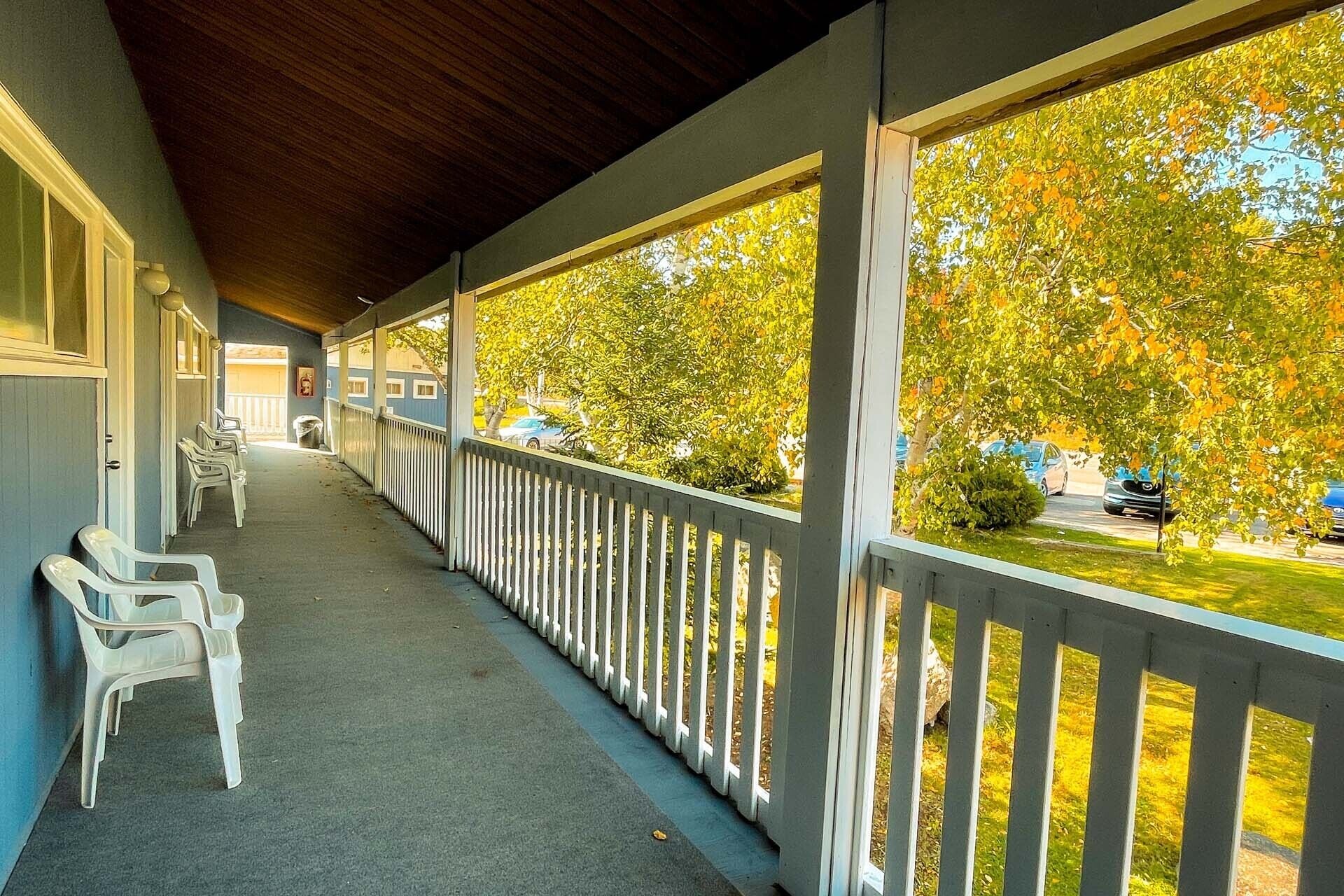 A covered porch with white plastic chairs, a railing, and a view of trees and parked cars.