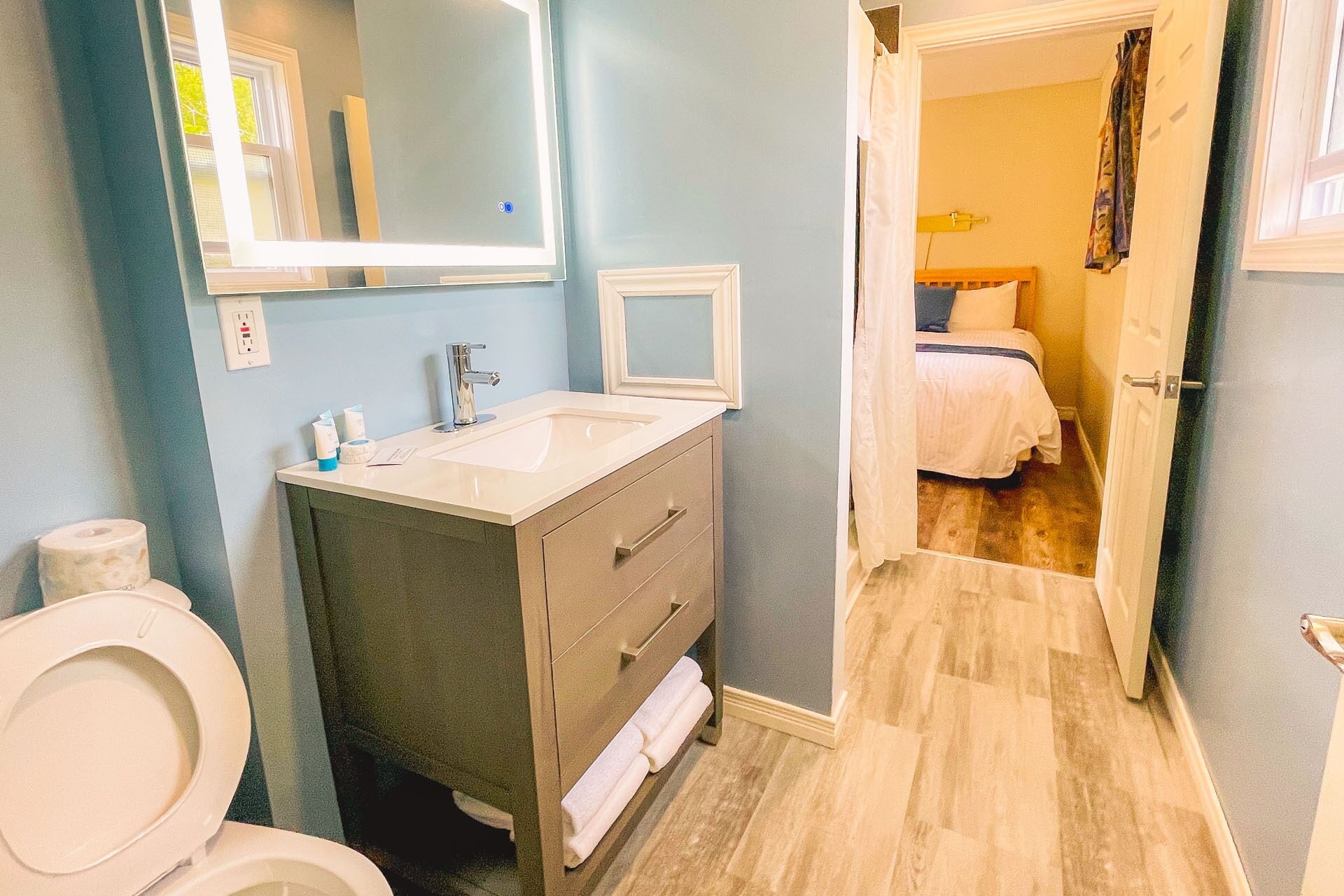 Un baño con inodoro, lavabo y espejo. Se ve una cama a través de una puerta abierta en la habitación contigua.