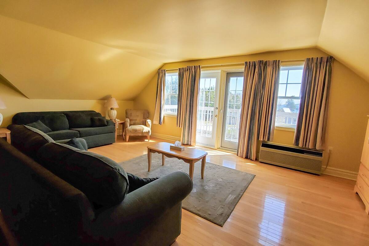 A living room with a dark green sofa, a wooden coffee table, and a patterned armchair. There is a sliding glass door with curtains, a small table with a lamp, and a radiator.