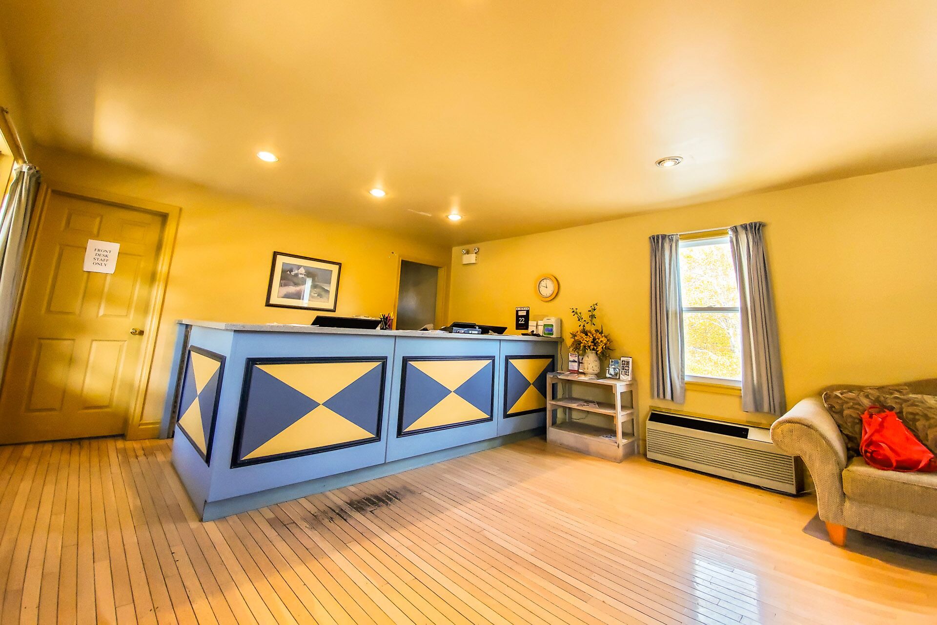 Una habitación con piso de madera, una recepción con diseño geométrico azul y amarillo, un reloj en la pared y una ventana con cortinas.