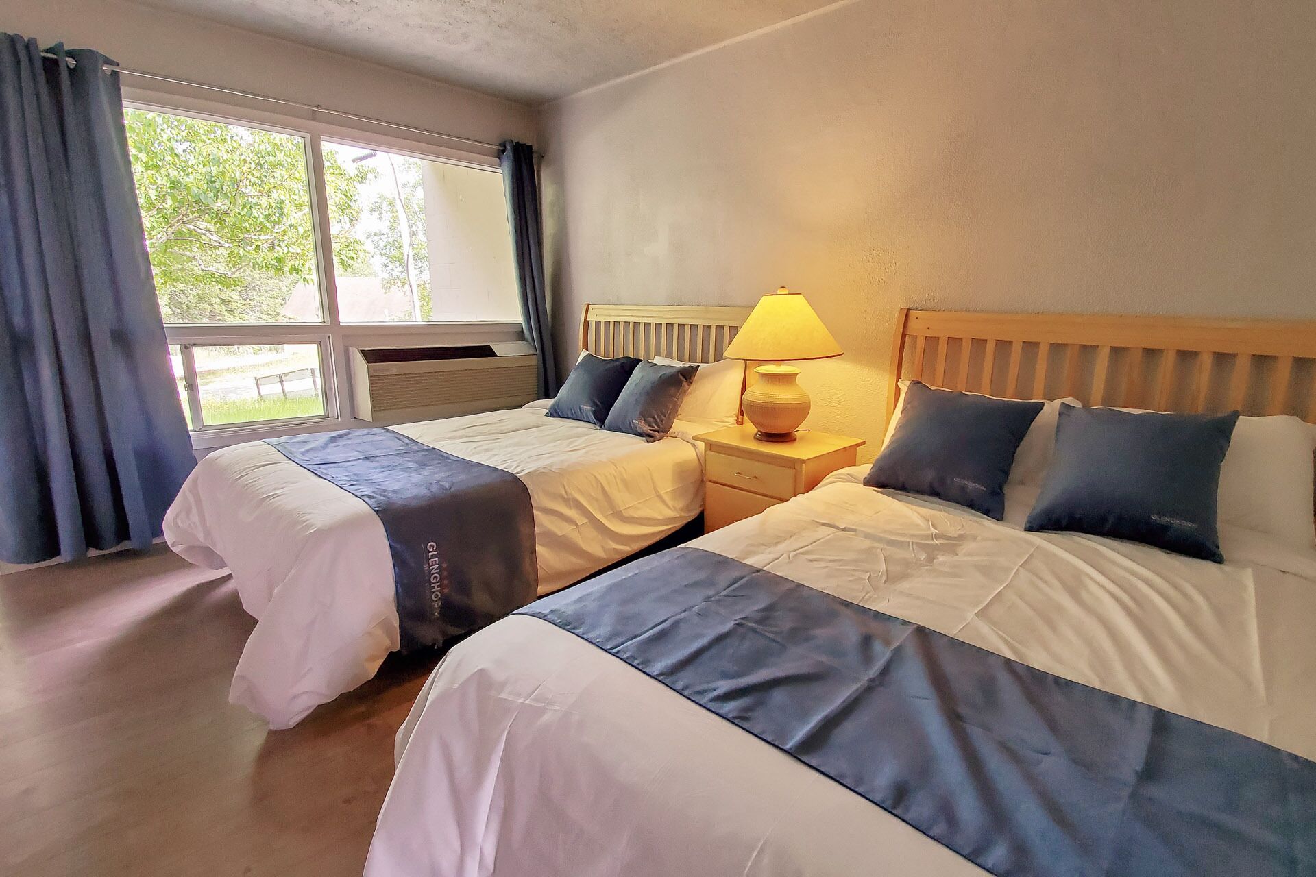 A hotel room with two beds, a nightstand with a lamp, and a window with curtains.