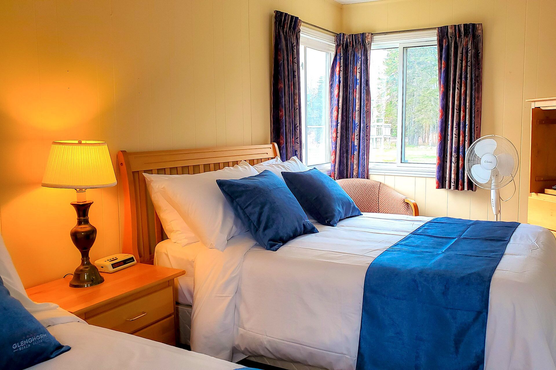 A hotel room with a bed, bedside table, lamp, and a fan.