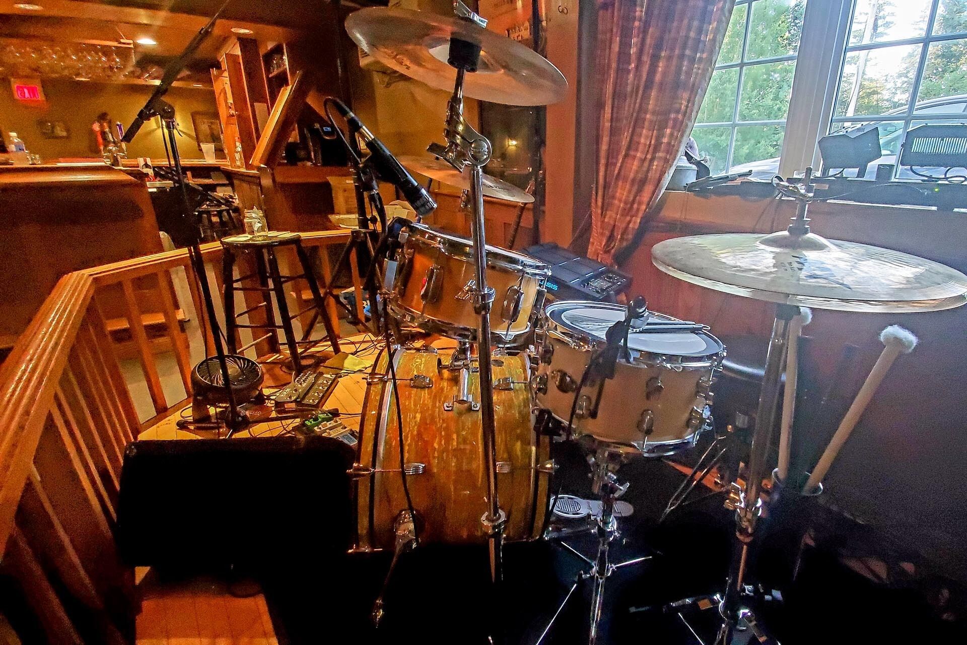 A drum set in a room with a window and a view of a car outside.