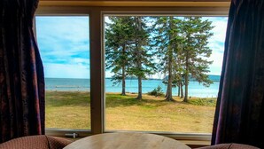A room with a large window offering a view of a grassy area, trees, and a body of water. - Glenghorm Beach Resort (Ingonish)