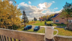 A wooden deck with a white mug, overlooking a residential area with two cars parked on the street, houses, and autumn trees. - Glenghorm Beach Resort (Ingonish)