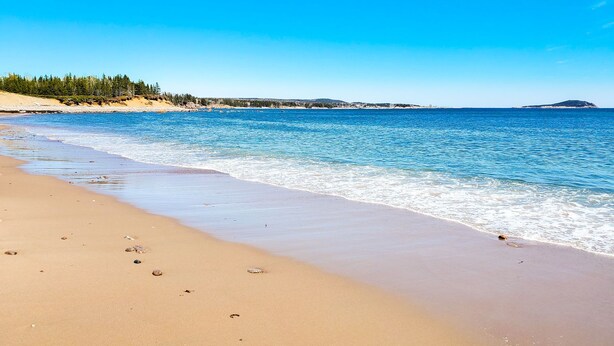 A beach with clear blue water, sandy shore, and a forested hill in the background. - Glenghorm Beach Resort (Ingonish)