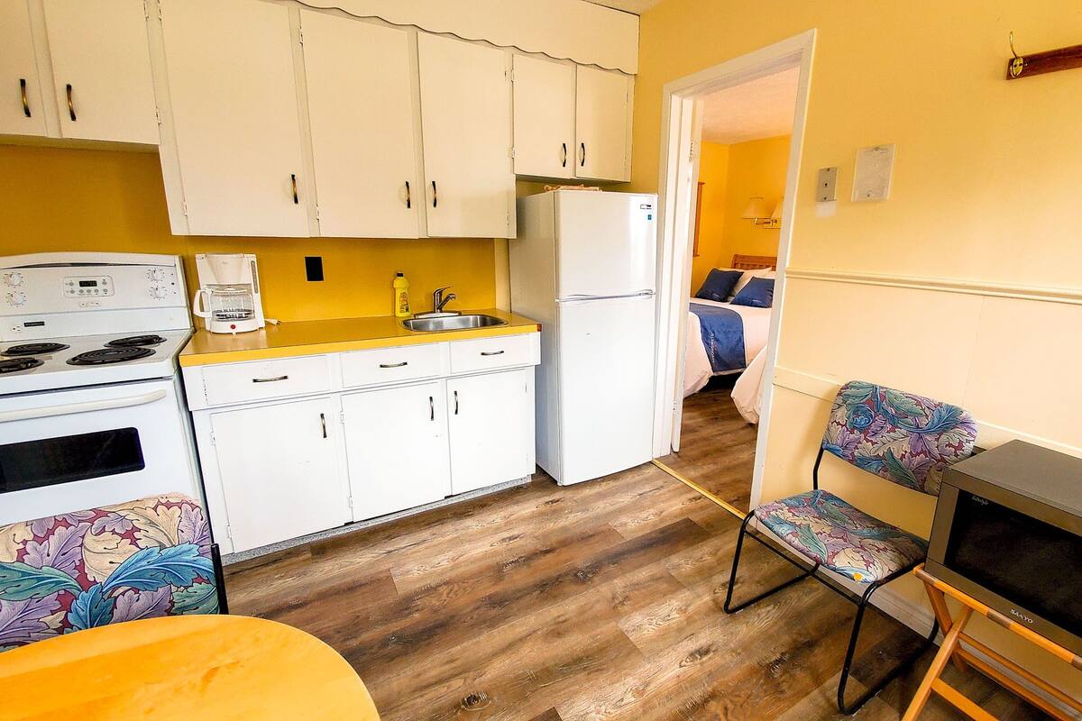 A compact kitchen with white cabinets, a stove, and a refrigerator. There's a wooden floor and a small dining area with a table and chairs.