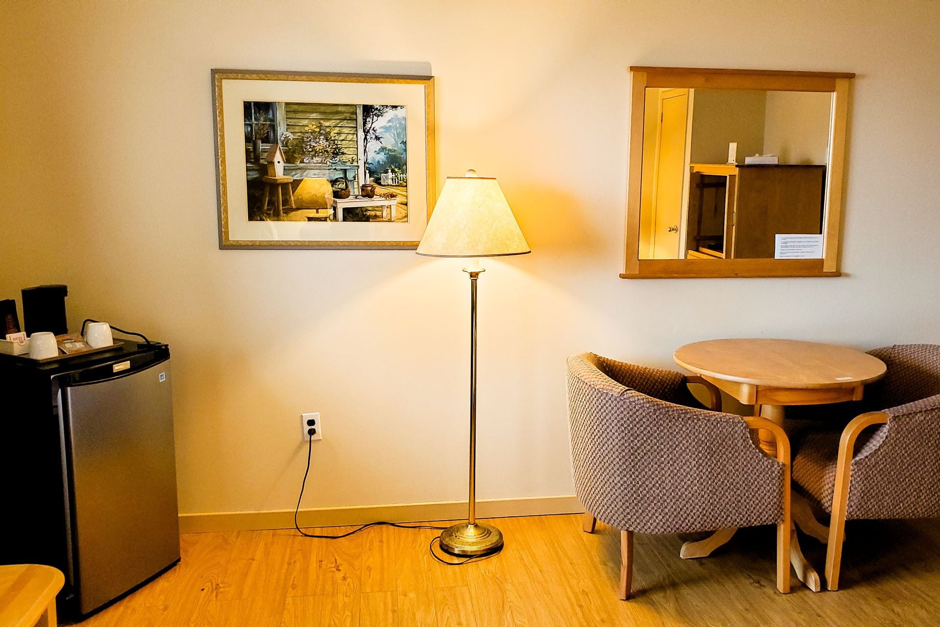 Une petite pièce avec un plancher en bois, une table avec deux chaises, un lampadaire, un réfrigérateur et un tableau encadré accroché au mur.