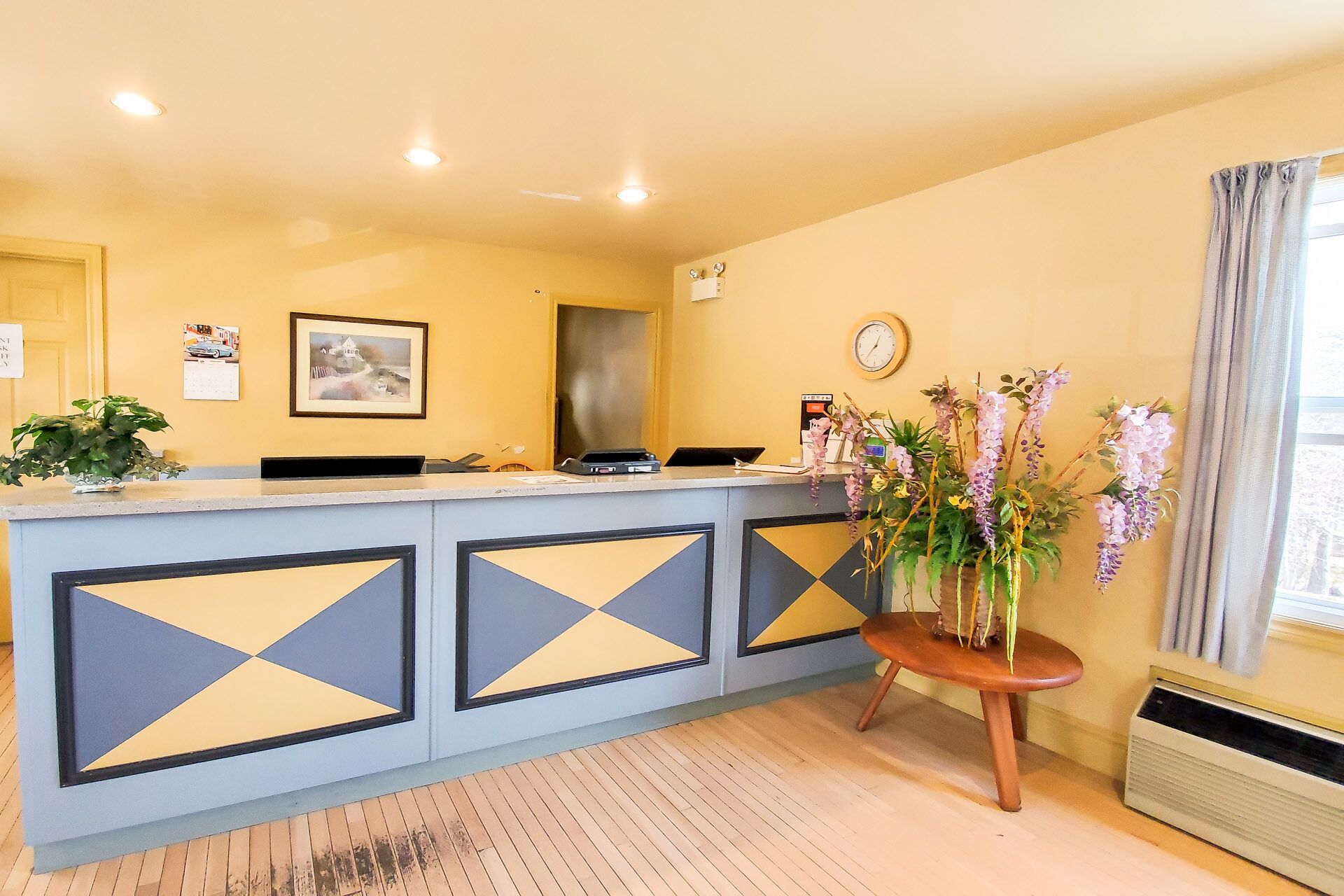 A reception desk with a modern design, featuring geometric patterns on the panels. There is a clock on the wall and a vase with flowers on a small table beside the desk.