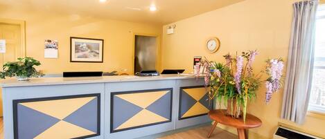 A reception desk with a modern design, featuring geometric patterns on the panels. There is a clock on the wall and a vase with flowers on a small table beside the desk.