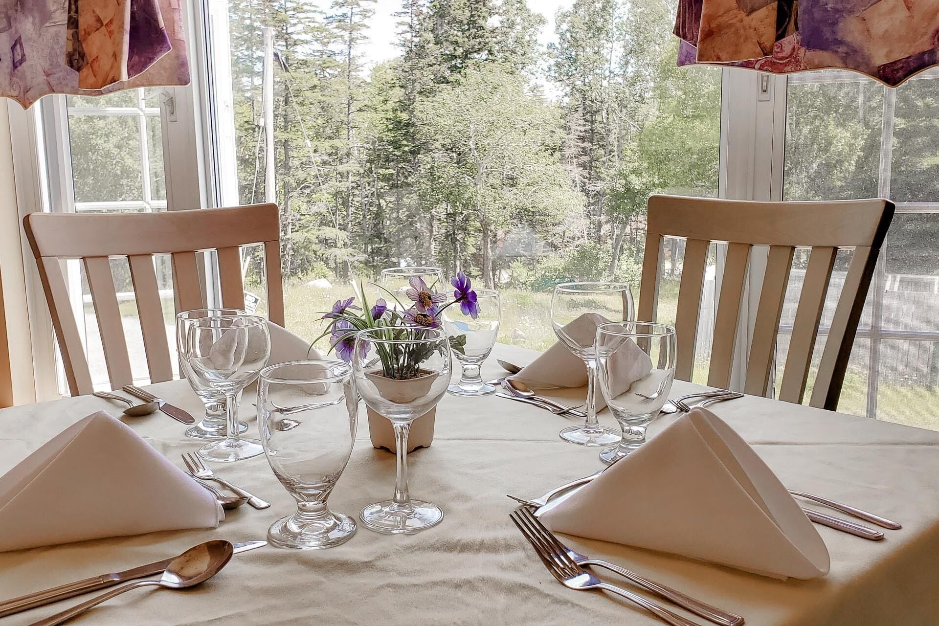Mesa de comedor para cuatro personas con sillas blancas, cristalería transparente y un pequeño jarrón con flores moradas.