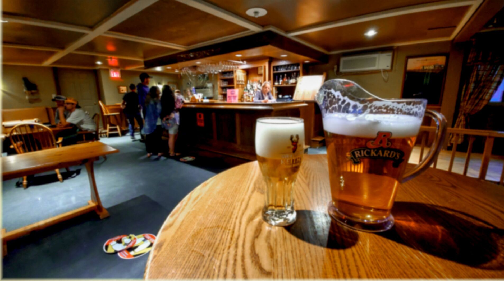 Un bar con mobiliario de madera, un vaso grande de cerveza y una jarra de cerveza sobre una mesa.