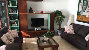 A living room with a brown sofa, a coffee table, a TV, a bookshelf, and a potted plant.