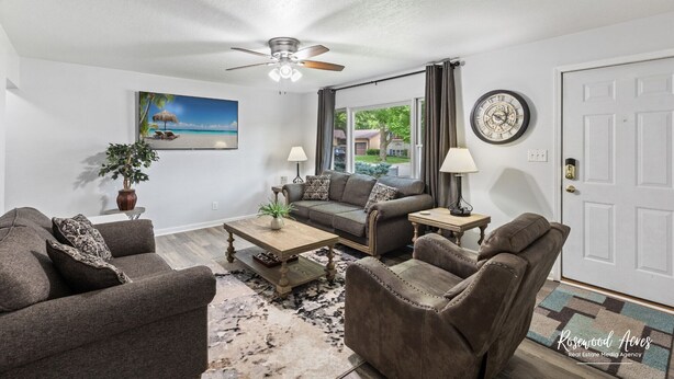 A living room with a ceiling fan, a painting of a beach, a sofa, a coffee table, and a clock on the wall.