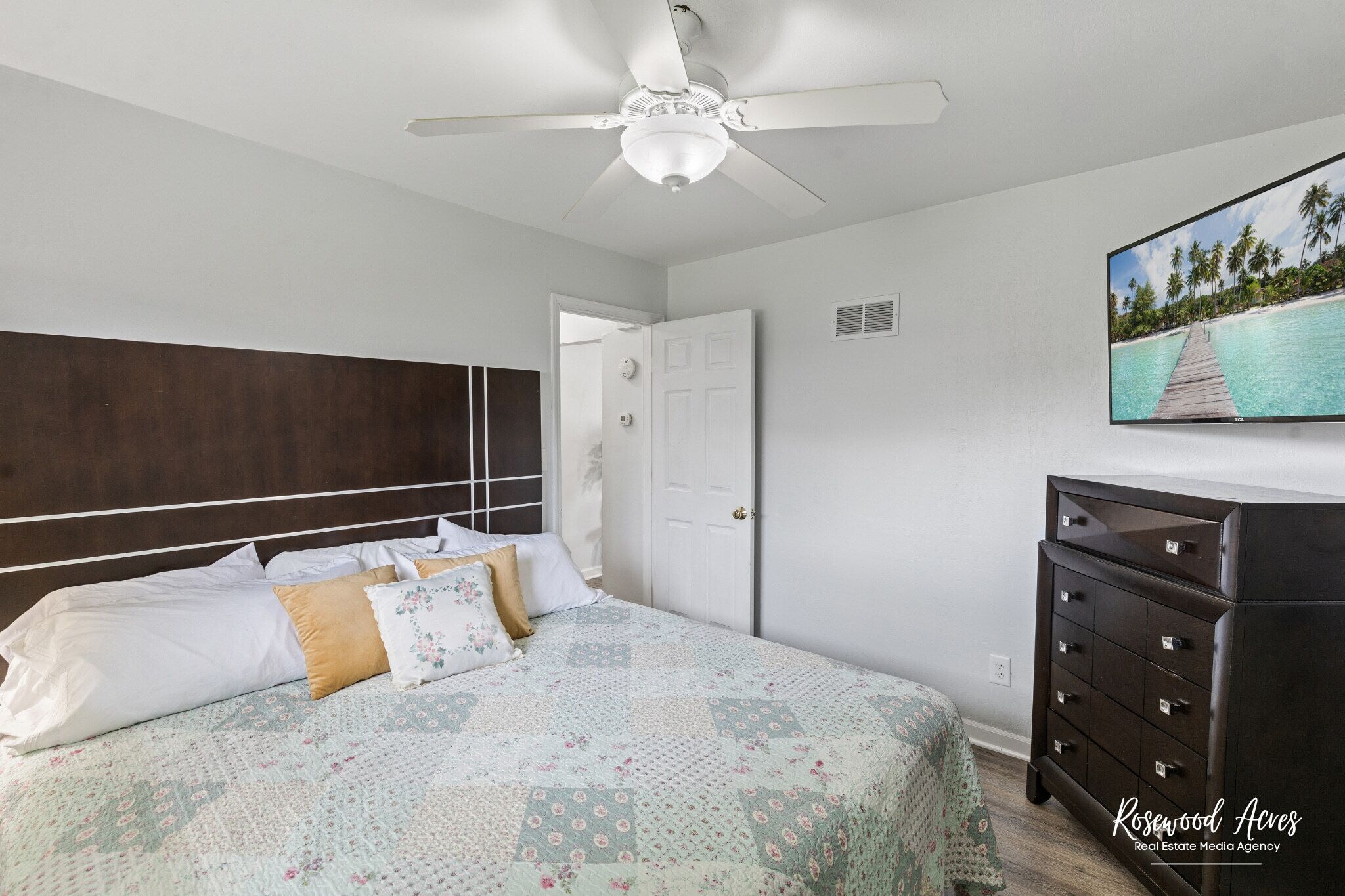 A bedroom with a bed, a dresser, a ceiling fan, and a wall-mounted TV.