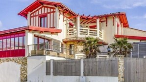 A two-story building with a red roof, white walls, and a stone base. There are two balconies with railings and a small garden area with palm trees. - Nice and spacious ground floor appartment close to the beach (LACANAU OCEAN)