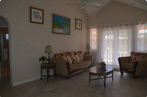 Un salon avec un canapé beige, une table basse ronde et deux fauteuils. On peut voir deux lampes sur des tables d’appoint, et un ventilateur de plafond est visible.