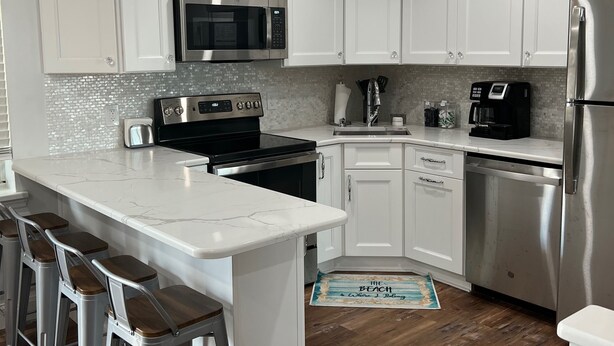 A modern kitchen with white cabinets, stainless steel appliances, a marble countertop, and wooden bar stools. - Avalon Stone Harbor - Heart of 7 Mile Island, Beach Block!! (Avalon)