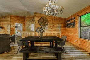 A wooden interior with a dining table, chairs, a chandelier, and a large window.