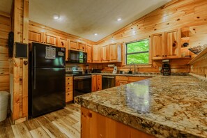 A kitchen with wooden cabinets, a granite countertop, a refrigerator, a microwave, and a window with a view of greenery.