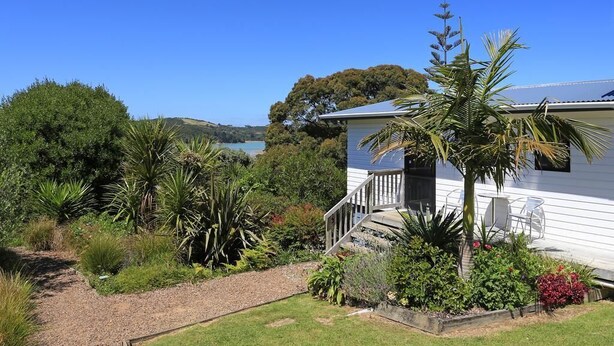 A white house with a covered porch, surrounded by a garden with various plants and a view of a body of water and hills in the background.