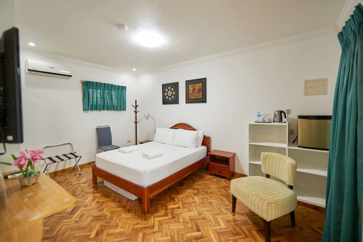 Een hotelkamer met een houten vloer, een bed met witte lakens, een bureau met stoel, een kleine tafel met een plant en een televisie.