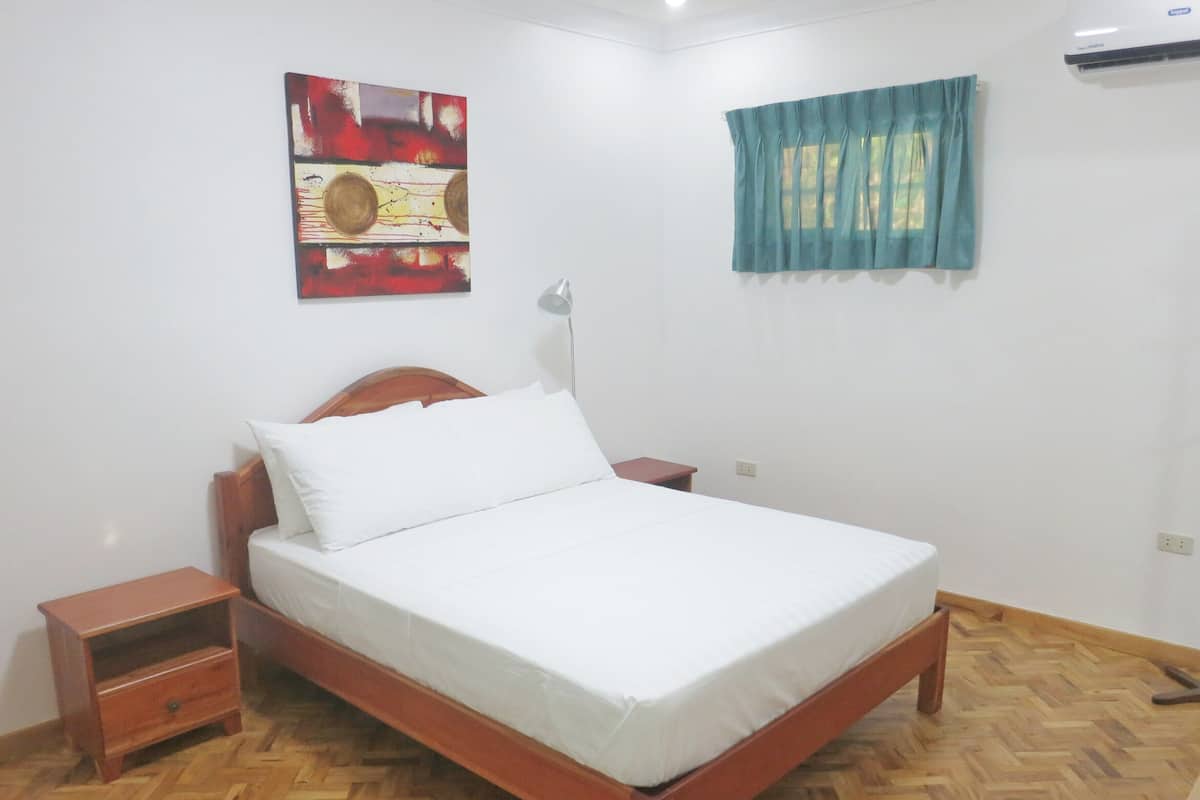 Een slaapkamer met een houten bed, nachtkastje, wandairconditioning, een raam met blauw gordijn en een schilderij aan de muur.