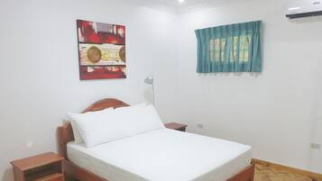 A bedroom with a wooden bed, a nightstand, a bedside table, a wall-mounted air conditioner, a window with a blue curtain, and a painting on the wall.