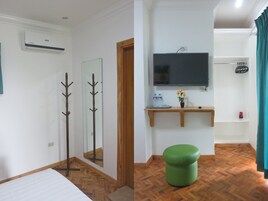 Habitación de hotel con cama, suelo de madera, reposapiés verde, espejo, perchero, televisor y estante con artículos de aseo.