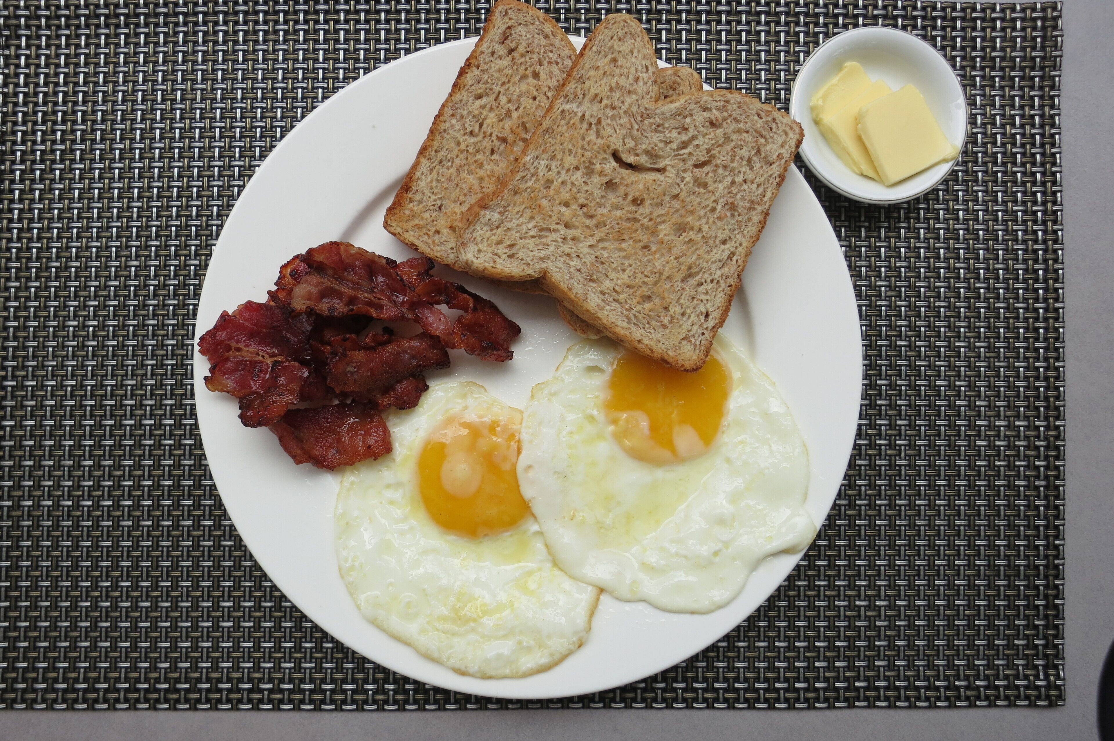 Un plato con tostadas, tocino y huevos fritos sobre un posavasos tejido.