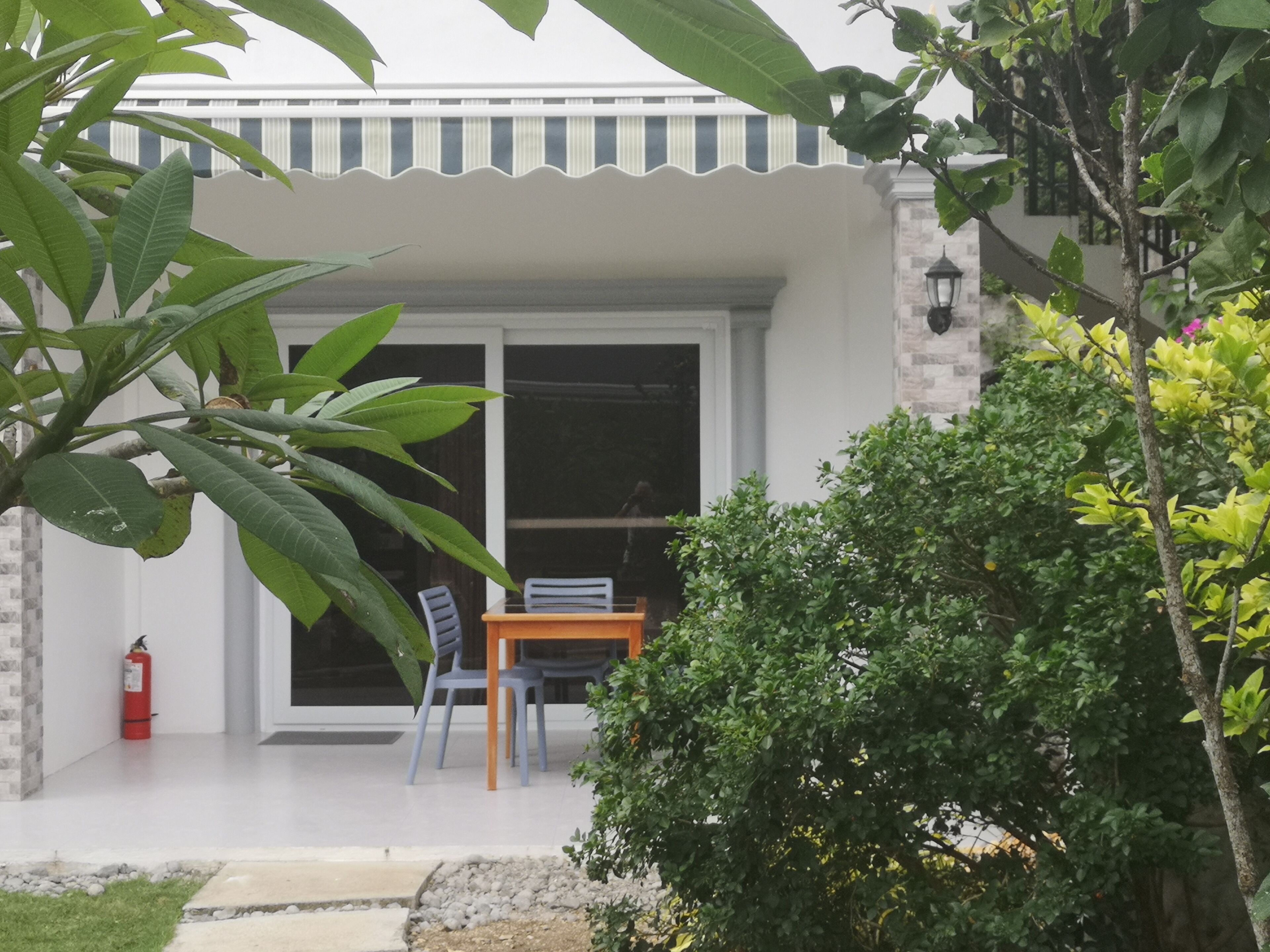 A covered outdoor seating area with a table and chairs, surrounded by greenery and a fire extinguisher.