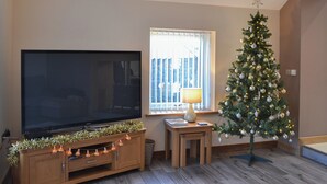 Una sala de estar con un televisor de pantalla plana grande sobre un mueble de madera, una mesita con una lámpara y un árbol de Navidad.