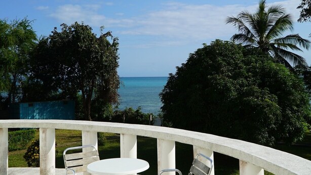 Terraço com mesa e cadeiras brancas, com vista para uma praia e palmeiras.
