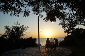 Vista sul tramonto da un'area esterna con posti a sedere in pietra, con sedie e tavolo in metallo, con vista su un corpo d'acqua.