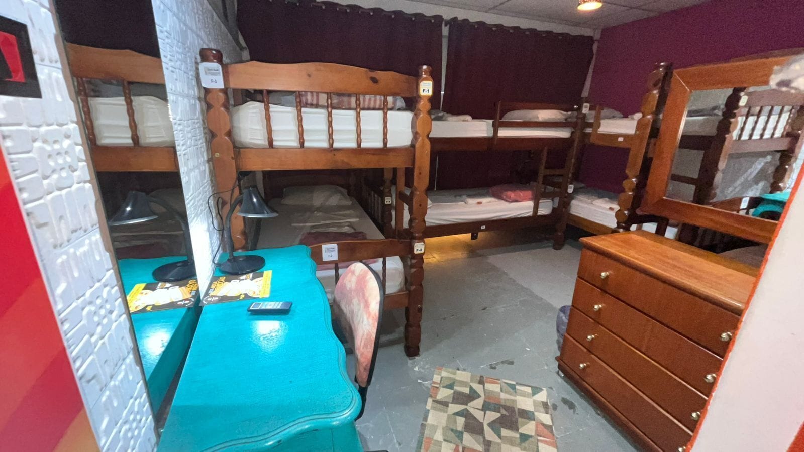 A dormitory room with bunk beds, a desk with a chair, a dresser, and a patterned rug on the floor.