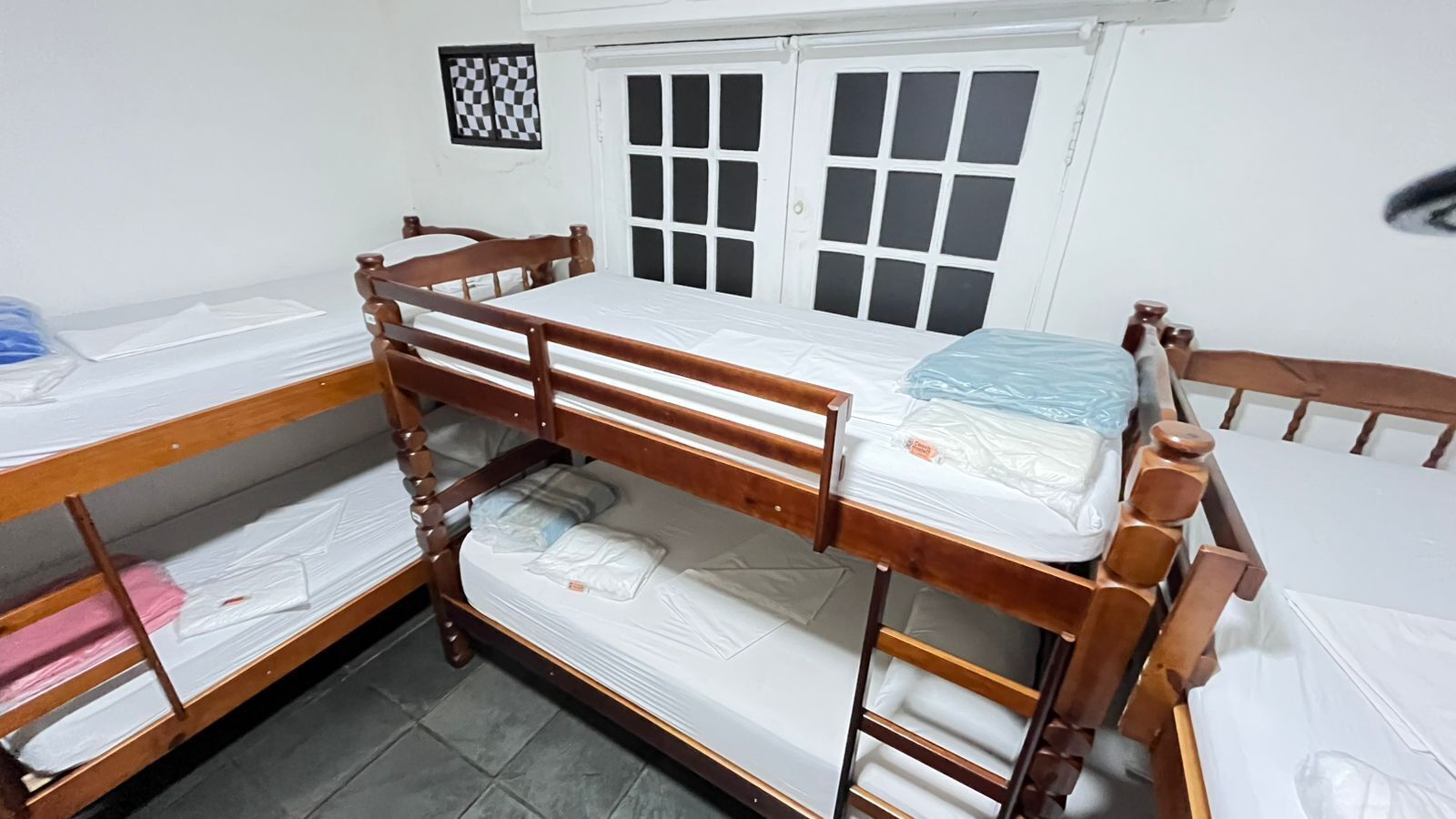 A dormitory room with bunk beds, a window with a checkered blind, and a door with frosted glass panels.