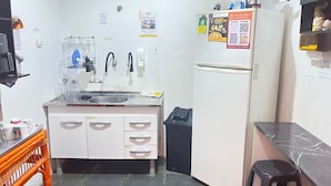 Uma cozinha compacta com pia, geladeira e lixeira. Há armários com puxadores e bancada com escorredor de louça.