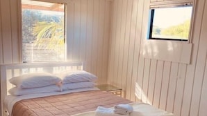 En säng med en brun sänggavel och vita sängkläder, ett litet sängbord, en hängande lampa och ett fönster med utsikt över grönområden.