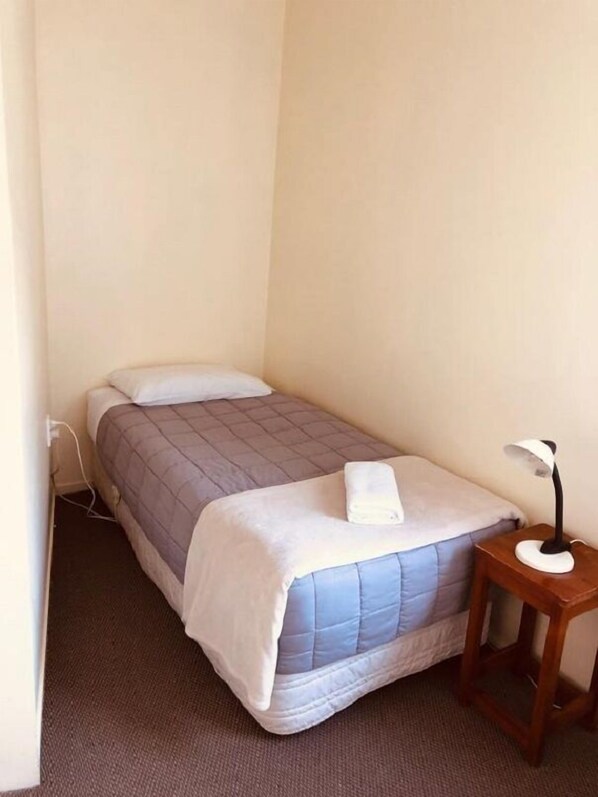 A small, single-bed room with a grey quilted bedspread, a white pillow, and a bedside table with a lamp.