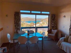 A room with a balcony, a dining table with chairs, a sofa, and a view of a lake and hills.