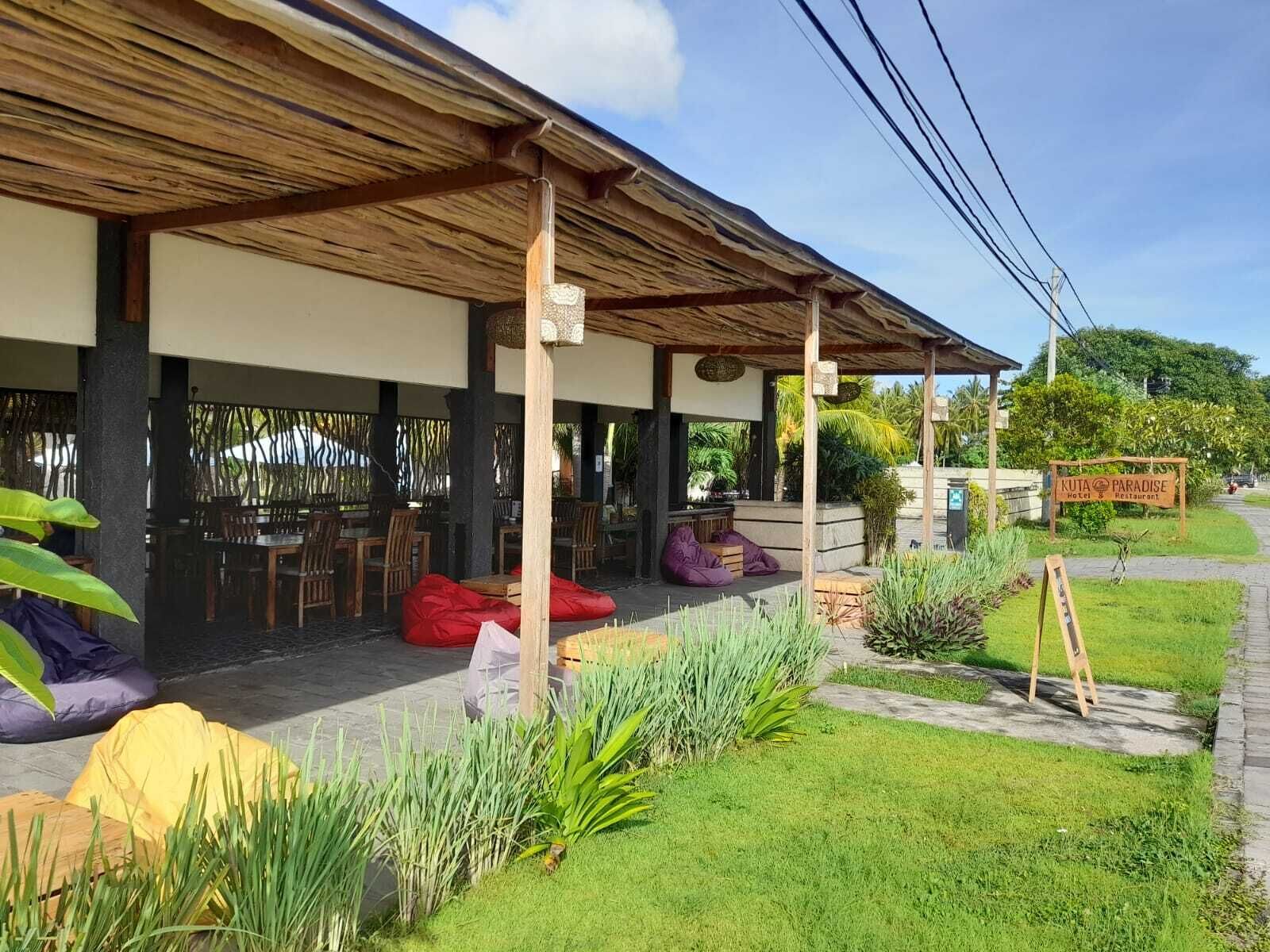 A covered outdoor seating area with wooden tables and chairs, surrounded by lush greenery and a sign for 'Kuta Paradise Hotel & Restaurant'.